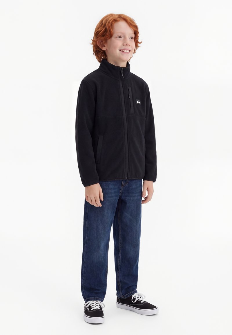 Garçon roux souriant debout, portant une veste noire zippée, un jean bleu et des baskets noires avec des lacets blancs sur fond blanc.