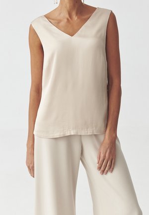 Vrouw draagt een beige mouwloze V-hals top en bijpassende wijde broek tegen een effen lichte achtergrond.