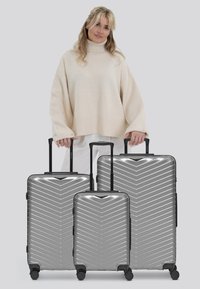 Trois valises rigides argentées de différentes tailles présentent un motif en chevrons, des poignées télescopiques noires et des roues lisses.