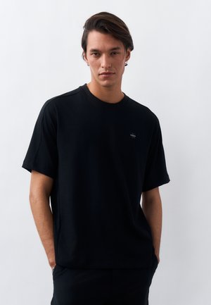 Νέος άνδρας με σκούρα μαλλιά που φοράει απλό μαύρο κοντομάνικο μπλουζάκι, στέκεται μπροστά σε λευκό φόντο με τα χέρια στις τσέπες.