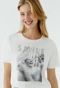 Wit T-shirt met een zwart-wit fotoprint van een lachende vrouw, versierd met strass-steentjes die boven de afbeelding het woord "SMILE" vormen.