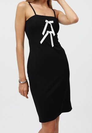 Femme portant une robe noire sans manches avec de fines bretelles et deux nœuds blancs sur la poitrine, posant avec un bras levé et un bracelet au poignet.