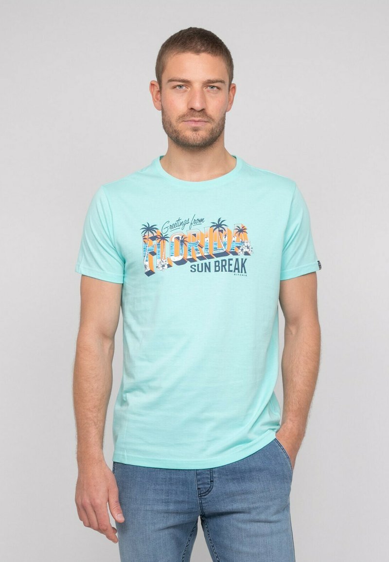 T-shirt vert menthe à manches courtes avec un imprimé graphique « Greetings from Florida » et des accents de palmier. Tissu doux et décontracté.