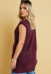 Överdimensionerad, ärmlös tröja i djup bordeauxstickad; har en ribbad nacke och sidofrånskärningar, bärs över ljusblå denimshorts.