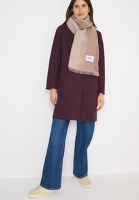 Viininpunainen takki, jossa on yksinkertainen muotoilu ja sivutaskut, yhdistettynä beigeen teksturoituun huiviin. Siniset levenevät farkut ja vaaleat lenkkarit täydentävät asun.