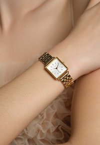 Reloj rectangular dorado con una esfera blanca, marcadores de hora dorados y una pulsera de eslabones metálicos curvados. Incluye una pantalla de fecha a las 3 en punto.
