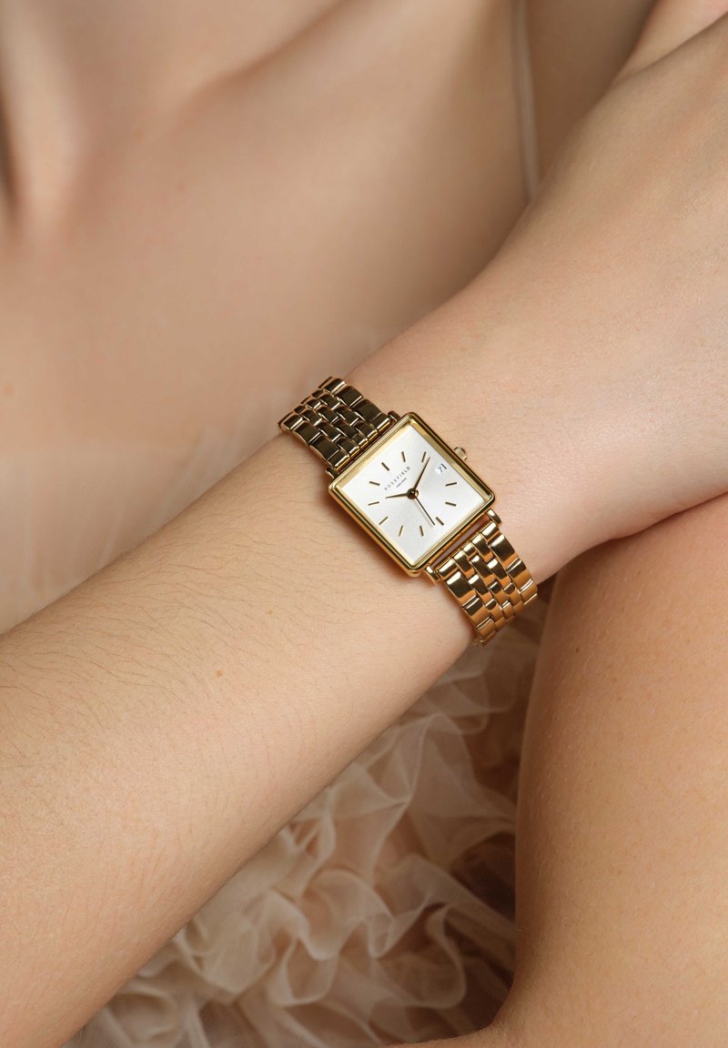 Reloj rectangular dorado con una esfera blanca, marcadores de hora dorados y una pulsera de eslabones metálicos curvados. Incluye una pantalla de fecha a las 3 en punto.