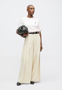 Modelis vilki baltą marškinėlį su mažai mėlyna grafika, smėlio spalvos plisuotas plačias kelnes, juodą diržą ir laiko juodą rankinę.