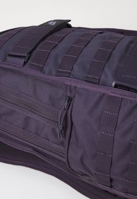 Saco de tecido roxo durável com alças de fita, bolso frontal com fecho e aba segura com um emblema. Design estruturado e compacto.