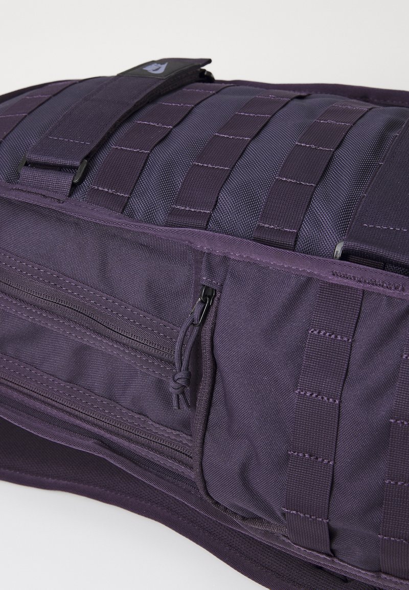 Sac en tissu violet durable avec des sangles en sangle, poche zippée à l'avant et un rabat sécurisé avec un badge logo. Design structuré et compact.