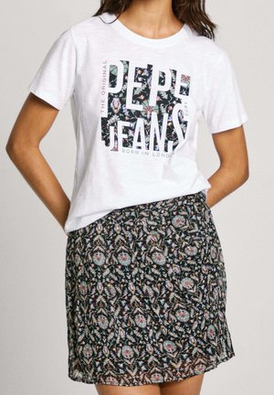 Nainen, jolla on valkoinen Pepe Jeans -t-paita kukikkaalla tekstillä ja tumma kukkakuvioinen hame, kädet lepäävät lanteilla.