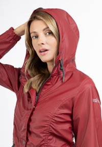 Jachetă waterproof roșie cu glugă, dotată cu închidere cu fermoar, manșete elastice și detalii plisate, confecționată dintr-un material neted și lucios.