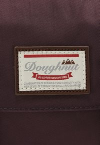 Etykieta marki pączków na ciemnym materiale z tekstem „Przygoda na świeżym powietrzu” i wyszywanym logo górskim na prostokątnej łatce.