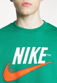 Ung person som bär en grön sweatshirt med en stor vit Nike-logotyp och en orange Nike-swoosh på bröstet.