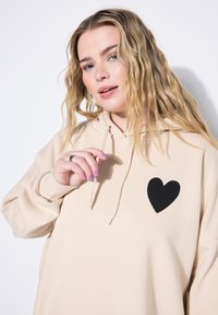 Sudadera beige con un gráfico de corazón negro en el lado izquierdo del pecho. Cuenta con capucha ajustable y textura suave. El modelo tiene el cabello ondulado.