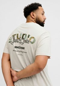 Jasnobeżowa bawełniana koszulka z nadrukiem na plecach. Z napisem „STUDIO Jack & Jones” w zielonym i czarnym kolorze oraz innym tekstem poniżej.