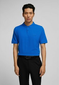 Polo bleu en tissu lisse, avec col, deux boutons et un logo discret sur la poitrine, associé à un pantalon noir et une ceinture.