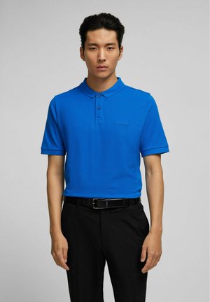 Blaues Polohemd aus glattem Stoff, mit Kragen, zwei Knöpfen und einem dezenten Logo auf der Brust, kombiniert mit schwarzen Hosen und einem Gürtel.