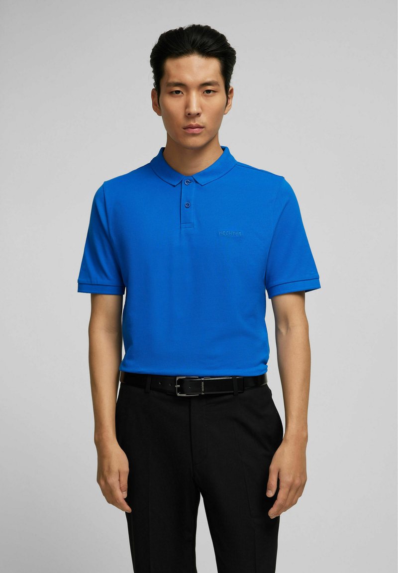 Polo bleu en tissu lisse, avec col, deux boutons et un logo discret sur la poitrine, associé à un pantalon noir et une ceinture.