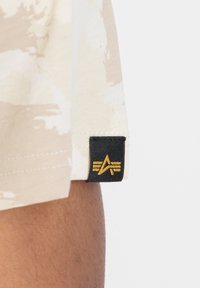 Beige mönstrad tyg med en svart etikett som har en stiliserad guldlogotyp. Etiketten är fäst vid fållen på ett plagg.