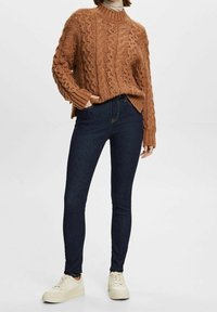 Suéter marrón de punto con un diseño de trenzas texturizadas, combinado con jeans oscuros ajustados y zapatillas blancas. Cuello alto con puños acanalados.