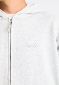 Svetlosivá zipová mikina vyrobená z mäkkého, štruktúrovaného materiálu. Obsahuje malé, razené logo na hrudi a strieborný zips s dvoma ťahacími pútkami.