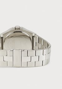 Sølv rustfritt stålklocke med en strukturert armbånd, som har et flatt oktogonalt hus, en rund bakside og en enkel krone øverst til høyre.