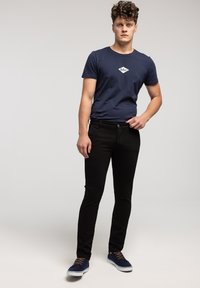 Granatowy t-shirt z białym logo, czarne dopasowane jeansy oraz granatowe sneakersy z brązowymi sznurowadłami, stojące na tle jednolitego szarego tła.