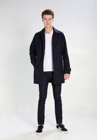 Navy coat with a button front, tailored design, and a collar, worn over a white shirt. Paired with dark jeans and black sneakers.