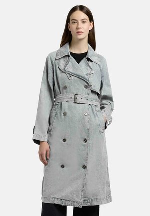 Kvinne iført en grå trenchcoat med belte, dobbelspent, brede slag og knappedetaljer, stående foran en ensfarget bakgrunn.