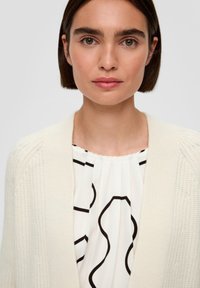 Romige geribde cardigan met een zachte textuur, gedragen over een witte blouse met zwarte abstracte patronen en geplooide details.