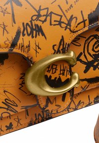 Sac à main en cuir fauve avec inscriptions noires style graffiti et une grande fermeture métallique dorée en forme de "C" sur le rabat avant.