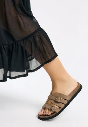 Sandalen aus Wildleder in Braun mit zwei verstellbaren Schnallen, einer konturierten Fußbettung und einer strukturierten schwarzen Gummisohle. Der Saum eines durchsichtigen schwarzen Kleides ist sichtbar.