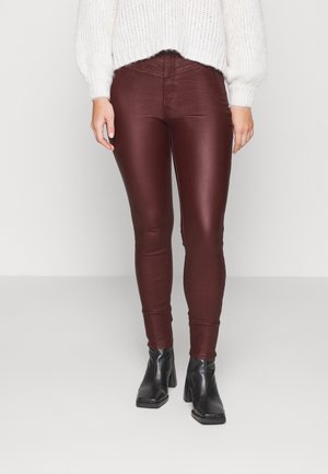 Femme portant des leggings en cuir rouge foncé, un pull en maille blanc et des bottines à talons noires sur un fond clair uni.