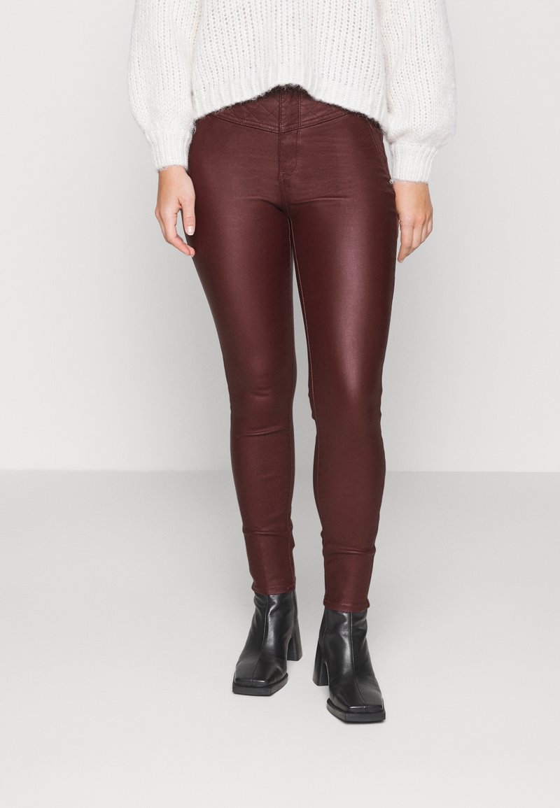 Femme portant des leggings en cuir rouge foncé, un pull en maille blanc et des bottines à talons noires sur un fond clair uni.