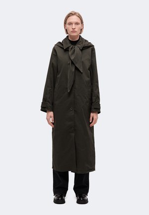 Une personne porte un long manteau vert olive foncé avec un col formé par une écharpe nouée, un pantalon noir et des chaussures noires, sur un fond clair et uni.
