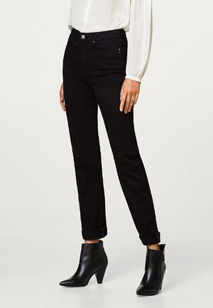 Kobieta w czarnych dopasowanych spodniach podwiniętych przy nogawkach, czarnych skórzanych botkach za kostkę oraz białej bluzce z długim rękawem, stojąca na białym tle.