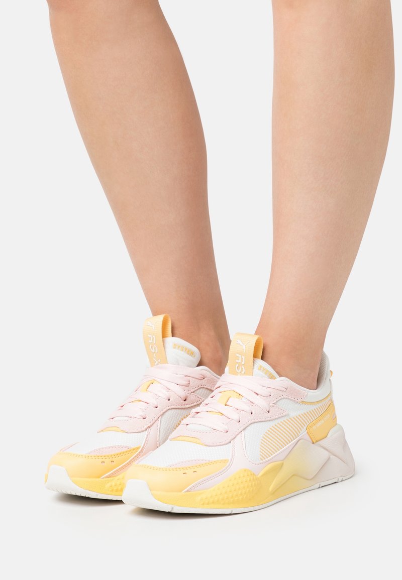 Pieds portant des baskets Puma RS-X jaune pastel et rose clair, debout sur une surface blanche avec un fond uni.