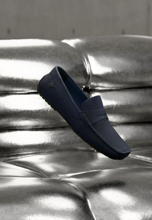 Zapato loafer azul marino flotando sobre un sofá de cuero plateado brillante con cojines capitoné en un entorno interior neutral.