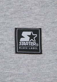 Grått stoff med en rektangulær svart etikett med hvit tekst som viser "STARTER" og en stjerne-logo, sydd på overflaten.