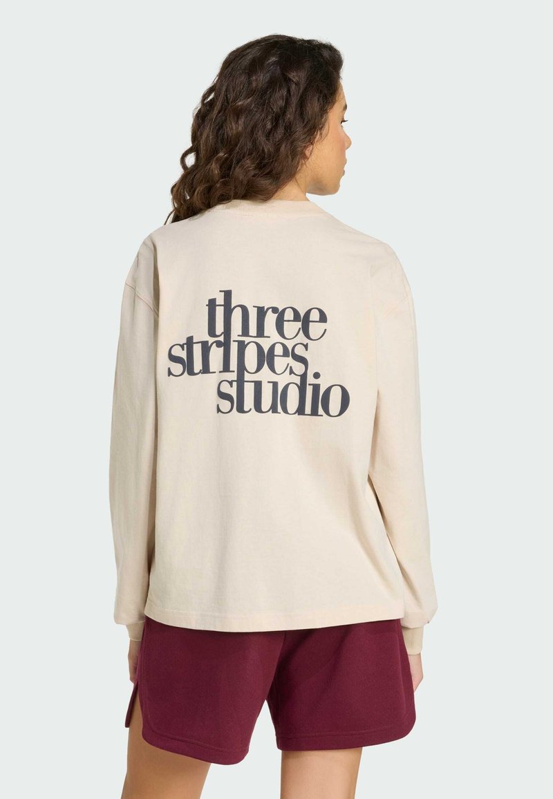 Beżowa koszula z długim rękawem z dużym czarnym napisem "three stripes studio" na plecach, zestawiona z bordowymi szortami. Gładka tekstura tkaniny.