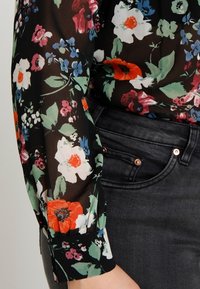 Blouse noire transparente à manches avec un imprimé floral coloré, associée à un jean en denim gris foncé montrant la poche avant et les rivets.