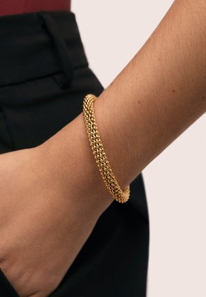 Pulsera de oro con acabado texturizado, con hebras entrelazadas y forma lisa y redondeada, colocada en una muñeca con ropa oscura.