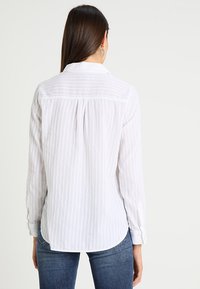 Kvinde med langt brunt hår iført en hvid langærmet stribet skjorte og blå jeans, vist fra ryggen mod en lys baggrund.