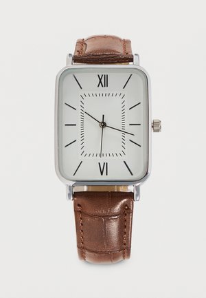Reloj de pulsera cuadrado plateado con esfera blanca, números romanos y marcadores negros, agujas negras y correa de cuero marrón con costuras.