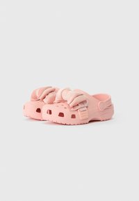 Clogs de Crocs em rosa claro com detalhes em veludo e babados na parte superior, buracos de ventilação e tiras ajustáveis no calcanhar, sobre um fundo simples.