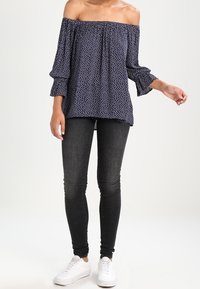 Kvinna klädd i en marinblå off-shoulder topp med vita prickar, svarta skinny jeans och vita sneakers mot en vit bakgrund.