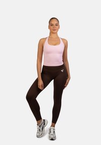 Jasnoróżowy top na plecach połączony z ciemnobrązowymi legginsami z wysokim stanem. Legginsy mają logo blisko pasa. Modelka nosi srebrne tenisówki.