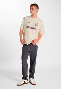 Lysebeige oversized t-shirt med SWARM-logo, kombineret med mørkegrå bukser og lyse sneakers med sorte detaljer.