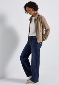 Veste beige avec un design à col, haut blanc, jeans ample bleu foncé et baskets de couleur claire. Les textures du tissu doux sont visibles.